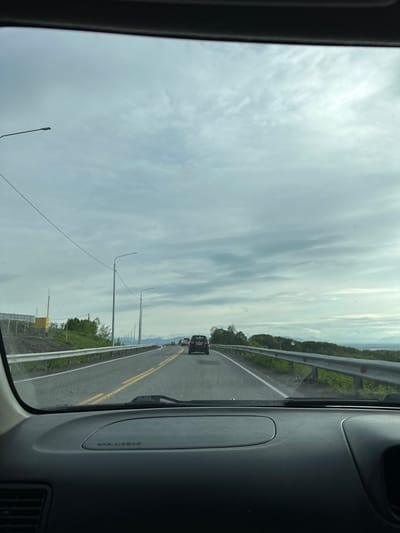 The image is a first-person perspective from inside a car, looking out through the windshield at a cloudy sky and a highway. The car is moving along a two-lane road with yellow dividing lines and a white edge line. On the left side of the road, there is a metal guardrail, a grassy embankment, and then what appears to be a construction site with a fence and a yellow structure. Several streetlights are visible along the road. Ahead, there are other cars on the road, including a dark-colored SUV. The landscape beyond the road is rolling hills covered in green vegetation, with distant mountains visible under the overcast sky. The dashboard of the car is visible in the foreground, with the text "SRS AIRBAG" imprinted on the passenger side. The scene suggests a drive through a somewhat rural or suburban area.
