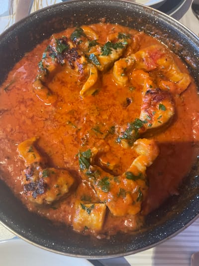 This is a close-up, top-down shot of a black, speckled pan filled with a creamy, orange sauce containing chunks of white fish and sprinkled with chopped green herbs. The fish pieces vary in size and shape, with some showing slightly seared edges. The sauce appears rich and thick, coating the fish generously. The overall impression is that of a hearty, flavorful dish, likely prepared or served recently. The background is blurred, but hints of a striped tablecloth and a white surface suggest a dining setting. The lighting appears to be indoor, with a warm cast, possibly from overhead lights. No people are visible in the frame. The context of Punta Ala, Italy, suggests this might be a local seafood preparation. There is no visible text in the image.