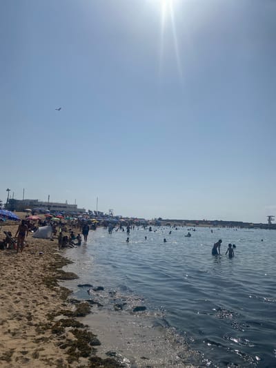 This is a full-frame, eye-level, outdoor shot of a crowded beach on a sunny day in Harhoura, Morocco. The image captures a bustling scene with a sandy beach on the left and the ocean on the right. Numerous people are scattered across the beach and in the water, enjoying the weather. On the sand, many beachgoers are seated under colorful umbrellas or relaxing on towels. Some are wading in the shallow water near the shore, while others are swimming further out. The ocean is calm with gentle waves, and the water is a deep blue. In the distance, along the coastline, there are buildings and structures, suggesting an urban or resort area. The sky is clear blue with the sun shining brightly, causing lens flare in the upper right portion of the image. A kite is visible in the sky in the upper left corner. The overall impression is one of a lively and popular beach destination.
