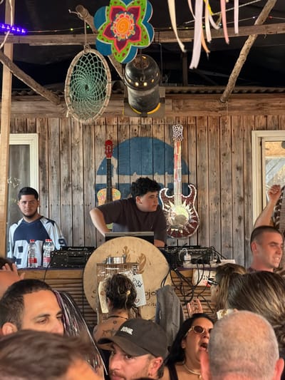 This image captures a lively outdoor or semi-outdoor social event in Pardes Hana - Karkur, Israel. The scene is dominated by a DJ booth constructed from rustic wooden planks. Two young men are positioned behind the DJ setup: one on the left, wearing a blue and white "99" jersey, stands idly with two water bottles on the Pioneer DJ controller in front of him; the other, center, with curly hair and a brown shirt, actively operates a laptop and DJ mixer. In front of the DJ, a large round object with a leaf design and "Pybx" text is visible.

The wooden plank wall behind the DJs is decorated with a blue abstract shape, a red acoustic guitar silhouette on the left, and a mosaic-style electric guitar silhouette on the right, featuring a central mirror. Electrical wires and power strips are visible behind the equipment. Overhead, rustic wooden beams support a dark canopy or roof, from which various decorations hang: a teal dream catcher, a colorful flower-shaped cutout, a black stage spotlight, purple string lights, and numerous white and pastel ribbons.

The foreground is filled with a dense crowd of attendees. Notable individuals include a man in a black "DONG." baseball cap looking towards the camera, a woman with sunglasses and her mouth open, and a man with short hair smiling and raising his right arm on the far right. Another woman, seen from behind, has dark hair, a necklace, and a visible Star of David tattoo on her upper back. The crowd's expressions and gestures suggest engagement with the music or event.