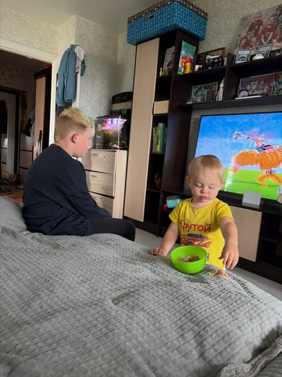 In a room with patterned wallpaper and a television screen displaying a cartoon, two young boys are present. One boy, with blonde hair, is seated on a grey, textured blanket on a bed, looking away from the camera. He is wearing a dark, long-sleeved shirt and dark pants. Beside him, a younger boy with blonde hair is also seated on the blanket. He is wearing a yellow t-shirt with a graphic of a red car and Cyrillic lettering. The younger boy is actively engaged with a green bowl containing food, with his hands reaching towards it. Scattered around the bowl are small pieces of food. The environment includes furniture such as a wardrobe, shelves filled with books and decorative items, and a fish tank. The scene appears to be an indoor domestic setting, likely a bedroom or living area, within the city of Centralny, Russia.