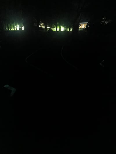 The image depicts an outdoor scene at night, possibly a park or recreational area, with artificial lights illuminating sections of the background and some faint lines on the ground. In the background, trees are silhouetted against lights, suggesting the presence of buildings or streetlights. The foreground is largely dark, with the exception of some faint, possibly painted, markings on the ground that appear to form curved lines and an angular shape. The overall impression is one of an empty, dimly lit public space at night.