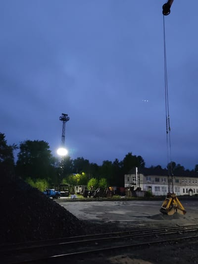 This image depicts an industrial or railway yard scene at dusk or night in Medvezhyegorsk, Russia. The sky is a deep twilight blue, with some visible cloud cover, and three small, bright points of light are discernible in the upper part of the sky.

In the foreground, multiple railway tracks run horizontally across the bottom of the frame. To the left, a large, dark mound of material, likely coal or gravel, dominates the lower-left quarter. In the mid-ground, a tall metal tower with a powerful floodlight stands prominently, casting a bright glow that illuminates the area and creates strong shadows. A blue truck or similar industrial vehicle is faintly visible behind the material pile, and other machinery can be seen in the distance near the floodlight.

On the right side of the image, the boom of a large crane extends downwards from the top, with cables leading to a substantial yellow grab bucket. The bucket is resting on the ground, holding a pile of lighter-colored material, possibly sand or gravel. In the background, an old, multi-story building with multiple windows and a somewhat weathered facade stands behind the crane and grab bucket, with trees surrounding the perimeter of the yard.

The scene suggests an active material handling operation, possibly related to railway transport or storage, occurring under artificial light during the evening or night. No people are visible, and there is no discernible text.