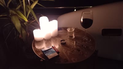 A low-angle, medium shot shows a table with three lit white pillar candles, a glass of red wine, a pack of "PARLIAMENT" cigarettes, a red lighter, a small metal grinder, a glass jar containing a granular substance, and a pipe with a metal bowl. The objects are arranged on a round table with a floral pattern. A potted plant with long, green leaves is visible to the left of the table. The background is dark, with a hint of a wall and a railing, suggesting an outdoor setting such as a balcony or terrace in Nerin, Montenegro, likely at night due to the presence of candles and the dim lighting. The scene is static, with no visible individuals or ongoing actions.