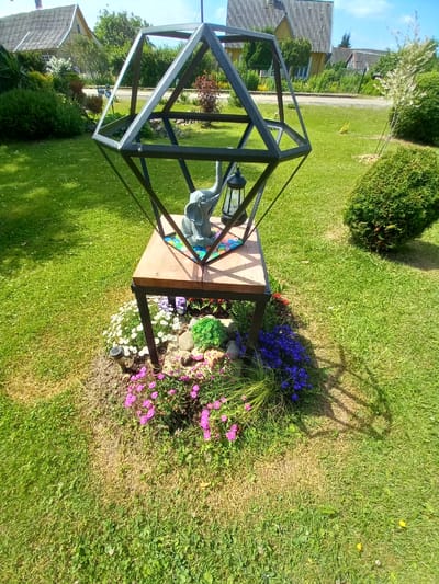 The image depicts a decorative garden feature in Adutiškis, Lithuania. The central element is a wooden table with a black metal frame, topped with a geometric metal structure resembling a geodesic dome. A grey elephant figurine and a black lantern sit on the wooden surface. The base of the feature is surrounded by a rock garden with various small flowers in shades of pink and purple, and some vibrant green foliage. A solar-powered garden light is also visible in the flower bed. The area surrounding the garden feature is a well-maintained lawn. In the background, typical Lithuanian houses with grey roofs and wooden facades are visible, along with trees and a clear blue sky. The overall scene suggests a sunny day in a residential area of Adutiškis.