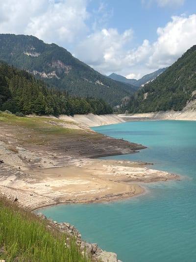 The image shows a view of a lake with turquoise water, surrounded by steep, forested mountains. The lake's edge is exposed in some areas, revealing a dry, terraced shoreline with remnants of tree stumps. The sky is partly cloudy with white clouds against a blue background. The location is identified as Lake of Sauris / Lago di Sauris in Lateis / Latais, Italy, a reservoir formed by a dam. There are no people or animals visible in the image. The environment appears to be a natural landscape with evidence of human intervention in the form of the dam and the exposed lake bed.
