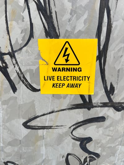 A yellow warning sign with a black lightning bolt symbol and the text "WARNING LIVE ELECTRICITY KEEP AWAY" is affixed to a metallic surface covered in black graffiti. The sign is torn and weathered. The metallic surface appears to be part of an outdoor utility box or enclosure. The graffiti consists of abstract black brushstrokes. The scene is likely an urban environment, possibly a street or alleyway in Balbriggan, Ireland, where such warning signs are common in areas with electrical infrastructure. No people are visible in the image.