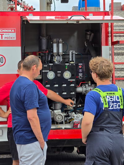 Two individuals are examining the controls of a fire truck. The person on the left wears a blue shirt and light grey athletic pants. The person on the right has curly blond hair, glasses and wears a blue shirt with the word "ZAHASICIO ŽEC" on it. The fire truck has the words "TECHNIKA PEČKY S.R.O." on its body. The fire truck controls include a gauge labeled "OTÁČKOMER" and a warning message "CHOO ČERP BEZ VODY MAX 60". The scene is in Mlčechvosty, Czechia.