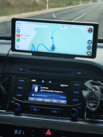 This media file captures an interior view of a car, likely during a road trip. The primary subjects are the car's dashboard and a mounted smartphone displaying a navigation app. The smartphone, positioned vertically, shows a map with a route highlighted in blue, indicating a journey in progress. The time displayed on the phone is 20:21, suggesting it's evening. The screen also shows signal strength (5G) and battery icons.
The car's central console features a car stereo system with various buttons and a rotary dial. Visible text on the stereo includes "DISP", "MUTE", "SCAN", "SETUP", "TUNE", "ENTER", "USB", "RADIO", "MEDIA", and "PHONE". The display also shows song information: "FILE 0009", "Home", and "Un Trandafir Creste La...". Other text elements on the stereo are "Info", "Shuffle", "Repeat", "Copy", and "List".
The background visible through the windshield appears to be a road or highway, with a blurred landscape of trees and a bright sky, characteristic of twilight. The overall scene suggests a person is driving or is a passenger in a car, navigating through an area. The car's interior is dimly lit, accentuating the illuminated screens. Location cues like "Sarmiziko", "3-ma", and "2-ga" on the navigation app might refer to street names or locations within Batovo, Bulgaria, as suggested by the context. No people are directly visible in the frame. The mood is functional and focused on the journey.