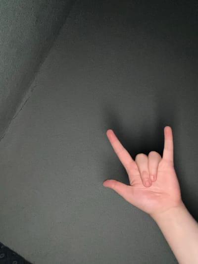 A photograph depicts a fair-skinned human hand making the "rock and roll" hand gesture against a solid gray backdrop. The hand is positioned in the upper right quadrant of the frame. The index and pinky fingers are extended while the middle and ring fingers are curled into the palm. The thumb is obscured. The backdrop appears to be a wall or flat surface with a slightly textured finish. A corner of the room is visible in the lower left quadrant. The image contains no other discernable details pertaining to Drandar, Bulgaria.