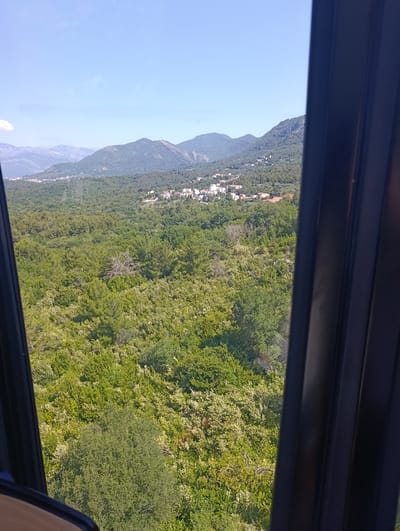 This media file is a photograph taken from a high vantage point, likely a cable car or a building, overlooking a densely forested hillside dotted with buildings. The scene is set in Kavač, Montenegro.

The foreground is partially obscured by dark, vertical framing elements, suggesting the viewpoint is through a window or opening. Beyond this frame, a vast expanse of lush green trees covers the landscape, with variations in texture and color indicating different types of foliage. Scattered amongst the trees, particularly on the slopes, are clusters of buildings with red-tiled roofs, forming small villages or residential areas.

In the background, rolling hills and mountains rise, their slopes also covered in vegetation. The sky is a clear, bright blue with a few wispy white clouds, indicating fair weather and a sunny day. The time of day appears to be midday or early afternoon due to the bright sunlight and shadows.

There are no people or specific objects of interest prominently featured within the landscape itself, apart from the natural elements and the man-made structures. The overall impression is one of a scenic, natural environment integrated with human settlement. The photograph captures a peaceful, pastoral view of the Montenegrin countryside. There is no visible text in the image.