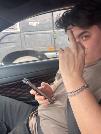 In this media file, a person is seen from a slightly elevated angle, looking towards the camera. Their right hand is raised, with a finger touching their nose. They are wearing a light brown t-shirt and dark grey pants. A silver chain bracelet is visible on their wrist. The person is holding a dark-colored electronic device, possibly a vape, in their right hand.

The background suggests they are inside a vehicle, looking out through the window. Through the window, a large truck or vehicle is visible, along with some other structures and markings that are difficult to decipher clearly. The interior of the vehicle shows black upholstery with red stitching.

The lighting suggests it might be daytime, but the exact time of day is not clear. There are no obvious indicators of weather conditions. The overall impression is a candid, informal shot, possibly taken while traveling.

The electronic device being held has "SAMSUNG" written on it.