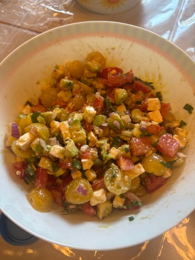 A close-up overhead shot shows a white bowl filled with a colorful salad. The salad appears to be a mixture of chopped tomatoes (red and yellow), avocado, red onion, and white cheese, possibly feta. Fresh herbs, likely parsley or cilantro, are also mixed in. The ingredients are coated in a light dressing. The bowl is resting on a wooden surface, and part of a clear plastic sheet is visible in the background, as well as a portion of a plate with a sun design. The overall lighting suggests an indoor setting. The image was taken in Merzenich, Germany.