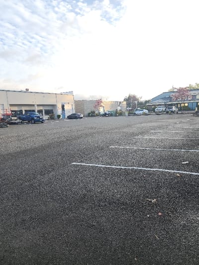 The scene is an asphalt parking lot in Lakewood, United States. The foreground displays dark gray asphalt with multiple white parking space lines and scattered small debris. In the midground, several vehicles are parked facing various commercial buildings. On the left, a blue Ford pickup truck is positioned near an orange scissor lift and industrial equipment. Directly behind these is a light-colored commercial building with multiple windows and a blue entrance door marked with the number "2097." To the right, a dark sedan, a light-colored sedan, and several white trucks or SUVs are parked in front of additional commercial structures, which include a lighter-colored concrete block building with reddish-foliage trees and another building featuring a blue sloped roof. The environment is an outdoor commercial area under an overcast sky, with no visible human presence or activity.