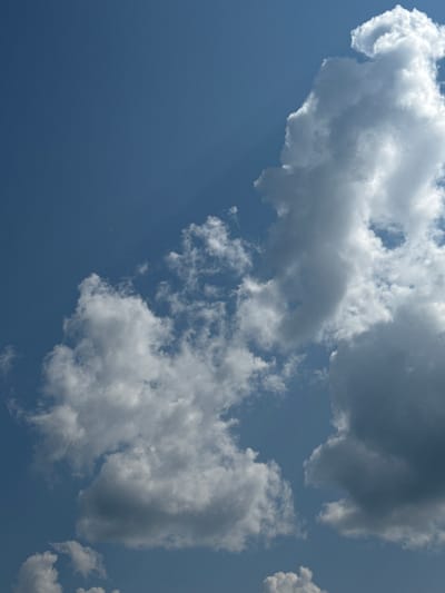 The image shows a bright blue sky filled with various types of clouds. In the upper right quadrant, there is a large, bright white cumulus cloud with distinct, fluffy edges, illuminated by sunlight. To the left of this cloud and lower down, there are several smaller, wispy clouds scattered across the sky. Towards the bottom of the image, more cumulus clouds are visible, some appearing darker and more dense, suggesting they might be further away or have more moisture. The lighting suggests it is daytime, likely midday or afternoon, given the brightness and the contrast of the clouds against the clear blue sky. There are no people, buildings, or any recognizable landmarks that would indicate a specific location within Chernoistochinsk, Russia or any other city. The scene is purely atmospheric.
