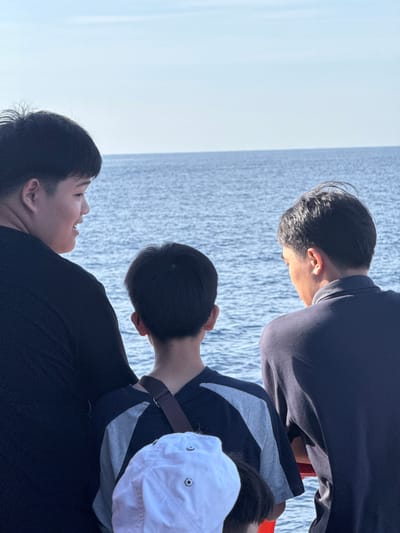 Three young males, viewed from behind, stand on what appears to be a boat or pier, looking out at the ocean under a clear sky. The male on the left, wearing a black t-shirt, is looking to his right, with his mouth open as if speaking. The middle male, in a gray and black shirt, has his head tilted down. The male on the right, in a dark blue polo shirt, also appears to be looking down. A portion of a white hat is visible in the foreground. The water is a calm blue, and the horizon is clearly defined against the sky. The scene is set outdoors, possibly in the city of Dong Son, Vietnam, given the presence of the ocean.