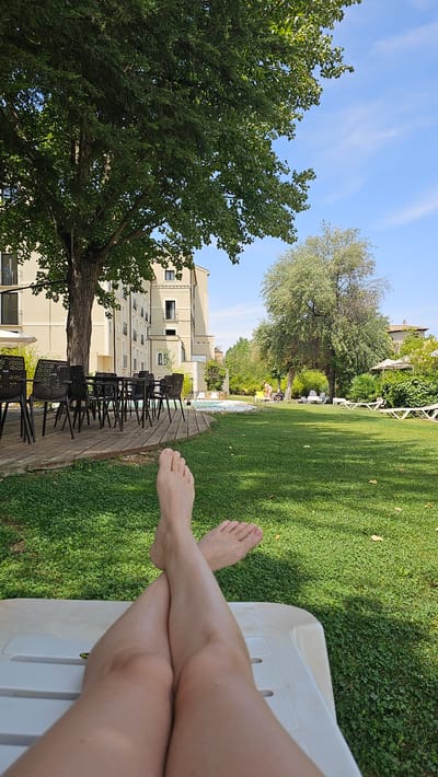 The image depicts a first-person perspective of someone relaxing on a white lounge chair in a sun-drenched outdoor area, likely a hotel or resort. The person's bare legs and feet are crossed at the ankles and are in the foreground, resting on the lounge chair. The background features a lush green lawn, a wooden deck with tables and chairs, a swimming pool, and a multi-story building. There are also several trees and some lounge chairs with umbrellas visible on the lawn. In the distance, there are people by the pool area. The sky is clear and blue with a few wispy clouds. The scene suggests a peaceful and leisurely atmosphere.