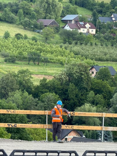 In the city of Jodłownik, Poland, a construction worker stands near a wooden fence. The worker is wearing a blue hard hat and an orange safety vest over a gray hoodie and dark pants. They are holding a mobile phone in their right hand and a pair of work gloves in their left. The fence has two horizontal wooden planks, each marked with "TLC" and "www.tlc.eu". Behind the fence, a wooded hillside is visible, with several houses and buildings nestled among the trees. The terrain is sloped, and the vegetation is lush and green, suggesting a rural or semi-rural setting. The weather appears to be overcast.