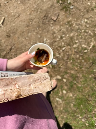 A downward-angled, outdoor shot shows a person holding a cup of tea and a book while standing on a dirt ground. The person is wearing a pink top and a pink sandal is visible. The white cup contains tea with what appears to be a tea bag and a few green leaves. The book is held in the same hand as the cup, with a visible spine printed with text in Russian, "Михаил Агеев. КАК МЫСЛИ СТАНОВЯТСЯ.." The ground is a mix of dirt, small rocks, and sparse patches of green grass or moss. The location is likely outdoors, but there is no detail in the background that defines it, nor places it in the city of Cheryomushki, Russia.