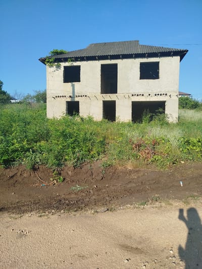 An unfinished, two-story concrete house stands in overgrown vegetation under a clear blue sky. The structure has a corrugated roof and empty window and door openings on both levels. Green vines creep over the left side of the roof. The ground in front of the house is muddy with tire tracks, transitioning to a gravelly path in the foreground. The shadow of a person holding a phone is visible on the path, suggesting the photo was taken by the viewer. The scene appears to be located in a rural or undeveloped area on the outskirts of Nimoreni, Moldova.
