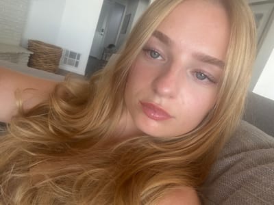 An image captured in Manhattan Beach, United States, depicts a close-up portrait of one individual, identified as a young female with long, wavy blonde hair and blue eyes, looking directly towards the camera. She has light skin, neutral facial expression, and appears to be wearing light-colored lip gloss. Her left shoulder is visible. The subject is positioned on what appears to be a gray, textured fabric sofa or cushion, which occupies the right foreground. The background, slightly out of focus, reveals an indoor residential setting. Visible elements include a light-colored brick or stone fireplace structure, a woven basket (possibly wicker), a white HVAC floor vent, and a doorway, suggesting a modern or contemporary interior. The lighting is bright and even.