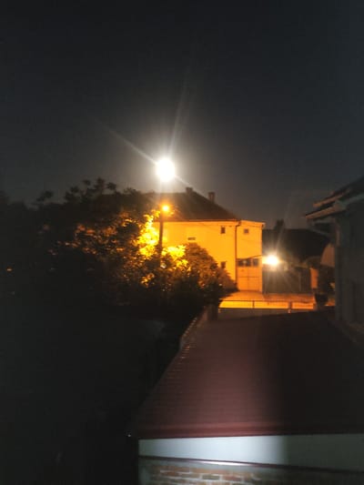 This is a nighttime photograph taken in Zrenjanin, Serbia. The image is oriented sideways and depicts an exterior scene. The primary subject is a building, appearing to be a multi-story structure with a bright yellow facade illuminated by artificial lights. One prominent light source, possibly a street lamp, is shining brightly on the left side of the image, creating lens flares and casting a glow on the surrounding darkness.
To the right of the building, a dense tree with illuminated foliage is visible, its leaves appearing golden or orange due to the artificial light. The background is a dark, hazy sky, suggesting it might be foggy or a very dark night. On the far right of the image, a section of a building with brickwork is visible.
The scene appears quiet and still, with no people or animals present. The activity, if any, is implied to be the general illumination of the area at night. Notable details include the strong artificial lighting creating stark contrasts and the almost abstract quality of the illuminated tree. The overall mood is somewhat mysterious due to the darkness and focused lighting. There is no visible text in the image.