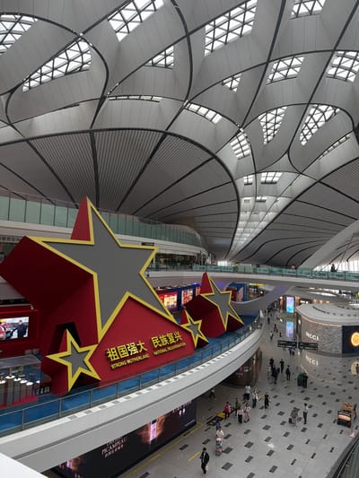 The image shows the interior of a large, modern building, likely an airport or a grand public space. The architecture features a distinctive, undulating ceiling with geometric skylights that allow natural light to enter. Dominating the foreground is a large, three-dimensional red star with a yellow outline, positioned on a raised platform. The star has a gray center and is adorned with Chinese characters and English text that reads "STRONG MOTHERLAND NATIONAL REJUVENATION." Below this display, a walkway or concourse is visible, populated by a number of people walking with luggage, suggesting a travel hub. Shops and storefronts, including one with a "GUCCI" logo, are visible on the lower levels. The overall environment is bright and spacious, with clean lines and a sense of grand scale. The location is identified as Guangyang District, China.