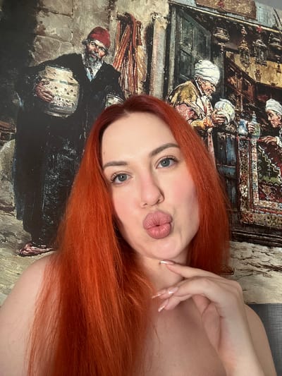 A fair-skinned woman with long, vibrant red hair is pictured in a close-up selfie, her lips pursed in a kissing gesture. She is positioned in front of a painting that depicts a scene with several men in traditional attire, set against a backdrop of what appears to be an outdoor market or bazaar. The painting features warm, earthy tones and intricate details, suggesting a historical or cultural theme. The woman's gaze is directed slightly off-camera, and her right hand is raised, with her index and middle fingers gently touching her chin. The background wall behind the painting has a subtle texture, possibly indicating an indoor setting in a residential area or an art gallery. The scene does not provide any specific visual cues to identify the location as Avcılar, Turkey.