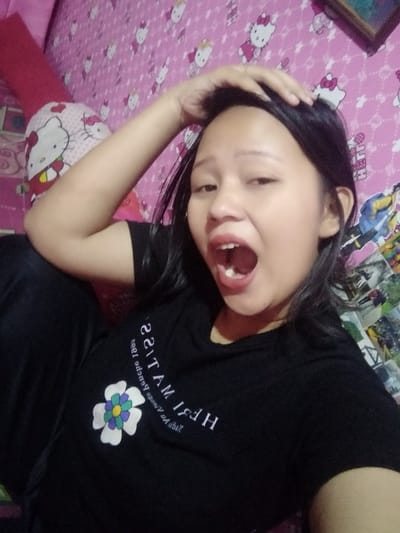 A young woman with dark hair and a black t-shirt adorned with a flower and reversed text poses for a selfie. She's lying on what appears to be a bed with a pink Hello Kitty-themed comforter and pillow. Her mouth is wide open in a possible yawn or shout, and her hand is resting on her head. The background includes a wall with more Hello Kitty patterns.