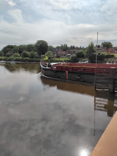 The image shows a large barge moving down a wide, calm river in Heiligenstedten, Germany. The barge is dark-hulled with a red upper rim and is creating a noticeable wake. Along the riverbank, there are several docked sailboats and a small residential area with houses and trees. The sky is partly cloudy, and the water reflects the sky and surrounding scenery. In the foreground, a portion of a wooden railing is visible, suggesting the image was taken from a bridge or pier.