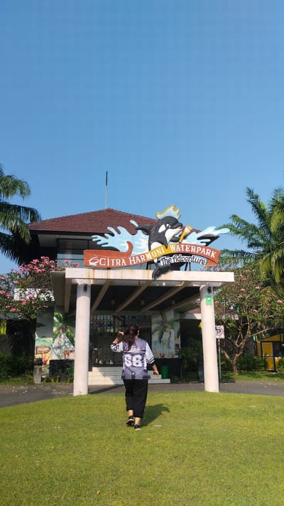 The image depicts the entrance to Citra Harmoni Waterpark in the city of Sidodadi, Indonesia. A woman, seen from behind, is walking away from the camera across a lush green lawn towards the entrance. She is wearing a dark jersey with white and grey accents and the number "88" on the back, paired with black pants. Above the entrance, a prominent sign features a colorful illustration of a killer whale leaping through waves, with the text "CITRA HARMONI WATERPARK The adventure" displayed. The entrance itself is supported by white pillars, and behind it, a building with a brown tiled roof is visible. The scene is set against a clear blue sky and surrounded by tropical foliage, including palm trees and flowering bushes. The overall impression is a bright, sunny day at an outdoor recreational facility.