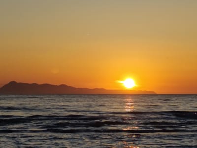 The image captures a serene sunset over the Aegean Sea, likely near Agios Stefanos, Greece. The sky transitions from a pale yellow at the top to a vibrant orange near the horizon. The sun, a bright, luminous orb, is positioned to the right of the frame, casting a shimmering reflection across the water's surface. In the distance, a silhouetted mountain range forms a dark, undulating line against the colorful sky. The sea itself is a deep blue, with gentle waves creating a textured, dynamic foreground. The overall atmosphere is one of tranquility and natural beauty. There are no people or man-made structures immediately visible in the scene.