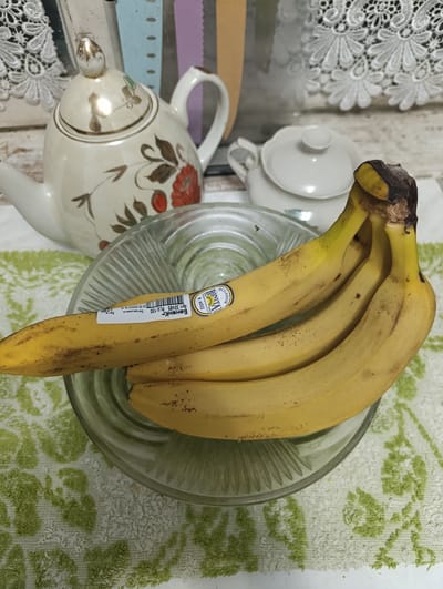A bunch of four ripe bananas rests in a clear glass bowl on a textured green and white cloth. The bananas are yellow with some brown spots, and one banana has a sticker labeled "Bonita" and "ECUADOR". In the background, a decorative teapot with floral motifs and a matching lid sits on a white surface, to the left of a smaller white sugar bowl. Behind these items, a patterned lace fabric is visible against a vertically striped background. The scene appears to be an indoor setting, likely a kitchen or dining area. The image has not been geolocated to Ladyzhyn, Ukraine.