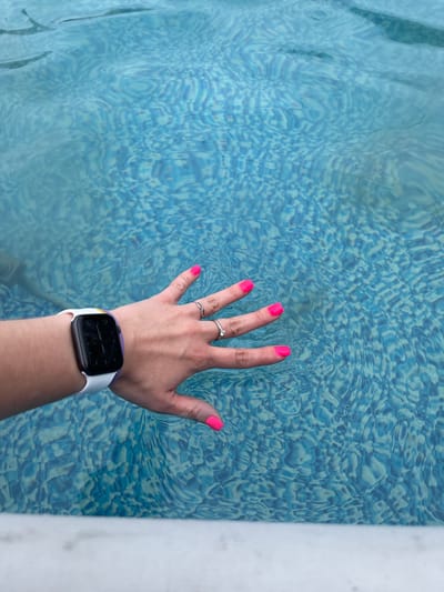 A person's left hand with bright pink fingernails and two rings is resting on the surface of a blue tiled swimming pool. A white smartwatch with a purple accent is visible on the person's wrist. The water is rippling, creating a distorted view of the pool's bottom. The scene appears to be in Plodopitomnik, Russia.