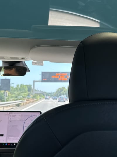 The image is taken from inside a vehicle on a highway, with the passenger seat in the foreground. A digital road sign is visible in the distance, displaying an orange text message that reads "ACCIDENT GRES SUR E NTIBES". Cars are driving on the highway in front of the vehicle. The weather appears to be clear and sunny. A GPS navigation system is displayed on a screen in the lower left of the image. The scene is likely in or near Villeneuve-Loubet, France, as indicated by the road sign mentioning "NTIBES" (Antibes), a city close to Villeneuve-Loubet.