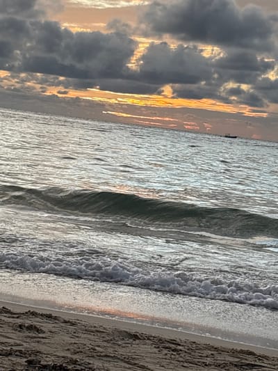 The image depicts a coastal scene during either sunrise or sunset in Roatán, Honduras. The foreground consists of light brown sand at the bottom of the frame, transitioning to the wet intertidal zone where a layer of white foamy surf from a breaking wave recedes. The midground shows the ocean with a series of gentle waves, including a more prominent, dark-crested wave just before it breaks. The water surface reflects the overhead sky. In the distant background, near the horizon and slightly to the right of the frame's center, a single small, dark vessel, consistent with a powerboat or panga, is visible on the water. The sky, occupying the upper two-thirds of the image, is characterized by extensive, dark grey cloud cover. Through breaks in these clouds and along the horizon, streaks of intense orange and yellow light are visible, indicating the presence of a low-angle sun. No individuals, discernible actions, or interactions are visible on the beach or in the immediate water.