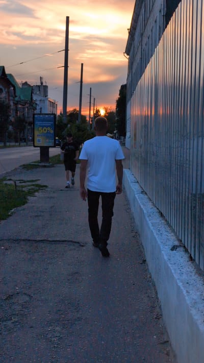 The image shows two individuals walking on a sidewalk during sunset in Starovirivka, Ukraine. The sky is filled with a dramatic orange and yellow sunset, with clouds catching the warm light. A corrugated metal fence runs along the left side of the frame, casting long shadows. The ground is paved with asphalt.
The foreground subject is a person wearing a white t-shirt and dark pants, walking away from the camera. Their posture is relaxed, suggesting a leisurely stroll. Behind them, another person in dark clothing is visible, also walking along the sidewalk.
In the background, trees and buildings are silhouetted against the vibrant sky. Streetlights are also visible, indicating a transition from daylight to evening. A digital advertisement panel is present on the sidewalk, displaying "50%" prominently, suggesting a sale or promotion. The overall mood is tranquil and serene, characteristic of a sunset scene. The time of day is clearly dusk.