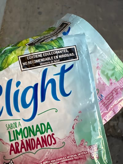 The image displays two flexible plastic pouches of a beverage mix, angled from an overhead perspective. The packages are predominantly light blue and green, with splashes of pinkish-red liquid depicted, suggesting the flavor. The product appears to be a diet or light version of a drink.

The package in the foreground is clearer and shows the brand name "light" in a large, stylized blue script. Below this, in green text, it states "SABOR A LIMONADA ARANDANOS," indicating a "Lemonade Cranberry" or "Lemonade Blueberry" flavor. The visual representation of the drink splashes is in a pinkish-red hue, suggesting cranberry.

A prominent black warning label with white text is visible on the front of both packages. It reads: "CONTIENE EDULCORANTES, NO RECOMENDABLE EN NIÑOS/AS. Ministerio de Salud." This translates to "CONTAINS SWEETENERS, NOT RECOMMENDED FOR CHILDREN. Ministry of Health."

Further details visible on the foreground package include "PESO NETO 80" (Net Weight 80) in the upper right corner, and smaller text describing the product as a "BEBIDA ANALCOHOLICA ARTIFICIAL DIETÉTICA" (Non-alcoholic artificial diet drink) and specifying "LIMONADA Y ARANDANOS" (Lemonade and Cranberries/Blueberries). Partially visible text on the upper package and other sections of the foreground one also mentions "LIBRE DE GLUTEN" (Gluten-free) and "CON VITAMINAS C Y D" (With Vitamins C and D).

The packages are glossy and appear to be sealed. The background is blurred and neutral, resembling a grey or concrete surface, providing little contextual information about the specific location or setting. The lighting is soft and even, highlighting the product packaging without harsh shadows. The items are stationary, suggesting they are either displayed, stored, or about to be consumed.