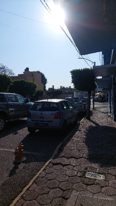 This image depicts a street scene in Tlaxcala City, Mexico, under direct sunlight from the upper right, causing lens flare and strong shadows. The street is asphalt with a yellow painted line marking the edge of a parking area. An orange, cone-shaped plastic traffic delineator with a white base rests on the asphalt in the lower-left foreground. Several vehicles are parked: a dark grey pickup truck is partially visible on the far left, followed by a silver Volkswagen Polo hatchback (MK4 generation) facing away from the camera. Behind the Polo, a white Nissan pickup truck is parked, displaying "NISSAN" on its tailgate. The sidewalk to the right consists of dark hexagonal pavers, with a metal utility cover embedded in the lower right. A small, trimmed tree stands on the sidewalk between the parked cars and a dark blue building facade, partially visible under a dark awning that extends into the upper right. In the background, other buildings are visible, including a light brown structure with a rooftop water tank on the left. The sky is clear and light blue. No individuals are clearly visible.