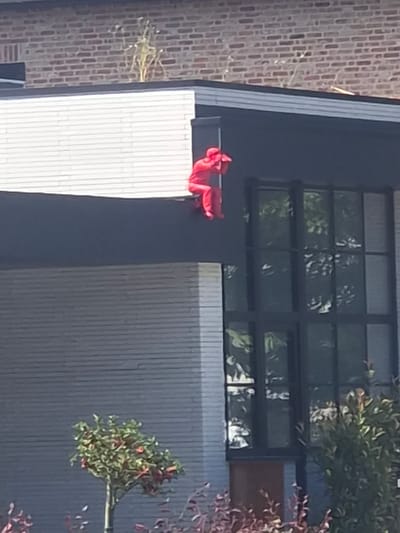 The image shows a person in a red outfit sitting on a ledge outside a modern building. The person appears to be facing away from the camera and is positioned on a dark, horizontal surface that runs along the exterior of the building. The building has a dark-framed, multi-paned window that reflects greenery, suggesting the presence of trees or foliage nearby. To the left of the person is a brick wall. To the right, there is a section of the building with a grey, ribbed facade and a small tree with green leaves and red berries. The overall scene suggests an outdoor urban or suburban setting, possibly during daylight hours given the illumination and reflections. No text is visible in the image.