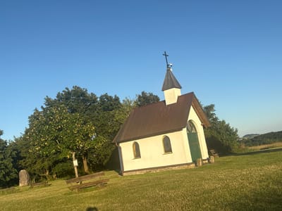 A small white chapel with a brown roof and a steeple topped with a cross stands on a grassy knoll under a clear blue sky. The chapel has a green door and two arched windows with yellow glass on its front facade. To the left of the chapel, there are trees and a wooden bench. To the right of the chapel, the ground slopes down to a landscape of rolling hills and trees. The scene is bathed in the warm light of the setting sun. This chapel is located in the municipality of Wershofen, Germany.