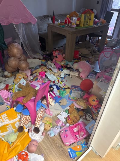 The image depicts a chaotic scene of a room overflowing with children's toys. A large wooden table, possibly a desk or play table, is a central element, with toys piled high around and on top of it. The floor is also covered in a multitude of stuffed animals, dolls, toy cars, balloons, and other playthings. A pink toy car and a pair of blue scissors are visible on the right side of the frame. In the lower right corner, a baby doll lies on its back amidst the clutter, partially obscured by an orange shopping bag with the text "MAKE YOU" and "COLORFUL BUBBLES" visible on other items. Several balloons with Cyrillic text, likely "Честит рожден ден!" (Happy Birthday!), are also present. The overall impression is one of a child's play area that has become overwhelmingly disorganized. The lighting suggests it could be daytime, but the interior setting provides no clear indication of the weather. The presence of numerous toys and a baby doll suggests a young child or children inhabit this space.