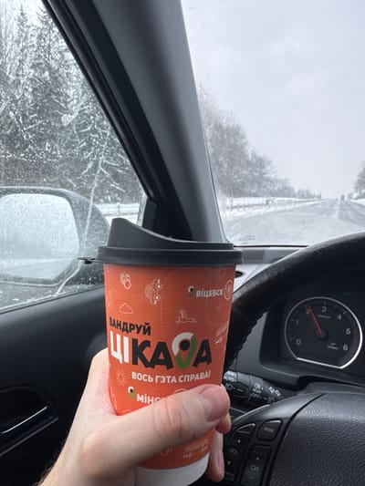 The image depicts a first-person perspective from inside a vehicle, capturing a winter scene in the city of Пяршайскі сельскі Савет, Belarus. In the immediate foreground, a fair-skinned hand holds an orange paper coffee cup with a black plastic lid. The cup is decorated with white stylized icons and Belarusian text, including "ВАНДРУЙ ЦІКАВА" (Travel Interestingly) and the city names "ВІЦЕБСК" (Vitebsk) and "МІНСК" (Minsk) with map pin graphics. To the right, a dark gray steering wheel and a tachometer are partially visible, with the needle pointing between 1 and 2, indicating approximately 1200-1500 RPM. The exterior environment, observed through the windshield, is snow-covered. A two-lane road, appearing wet or slushy from melted snow, extends into the distance, flanked by snow-dusted trees and a fence line on the right. The sky is uniformly overcast and bright gray. The vehicle's left side mirror and window display ice or condensation, partially obscuring the view of snow-laden trees beyond.