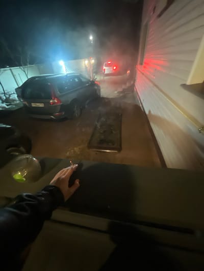 The image depicts a nighttime outdoor scene in Заозерное Снт, Russia. In the lower-left foreground, a human hand holds a lit cigarette, resting on a dark, flat surface next to a partially visible clear plastic bottle with a green cap. A dark grey station wagon, bearing Russian license plate T996AX 178, is parked in the midground, facing away from the viewer; its rear hatch appears partially open or damaged. To its right, a rectangular metal grate lies flat on the ground. Further in the background, a white van with illuminated red taillights is visible in a narrow passage, with ambient haze or smoke present in the air. The right side of the frame is occupied by the white-paneled exterior wall of a building, reflecting red light. Sparse, bare vegetation is visible on the far left.