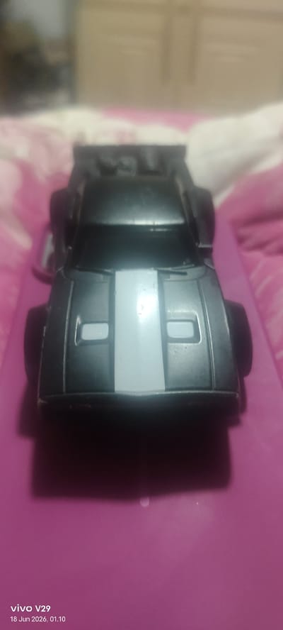 A close-up, slightly blurry, overhead shot of a black toy car with a silver stripe down the center of its hood, placed on a bright pink surface. The car appears to be a generic, stylized model with large, black wheels. The background is out of focus, showing hints of what might be bedding or fabric in muted colors. The timestamp "18 Jun 2026, 01.10" and "vivo V29" are visible in the lower left corner, suggesting the image was taken with a vivo V29 phone on that date and time. There is no indication of a specific location in Pulodolang, Indonesia within the image itself.