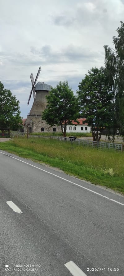 The image shows a historic windmill in Adavere, Estonia, on a cloudy day. The windmill, made of stone and with a metal roof, has four visible blades. In front of the windmill is a white building with a red roof and several windows. A metal fence separates the buildings and windmill from a grassy area with tall grass. A paved road with a white line runs across the foreground of the image. Trees are visible on both sides of the windmill and building. The bottom of the image contains text indicating the device used and the date and time the photo was taken: "REDMI NOTE 9 PRO AI QUAD CAMERA 2026/7/1 16:55".