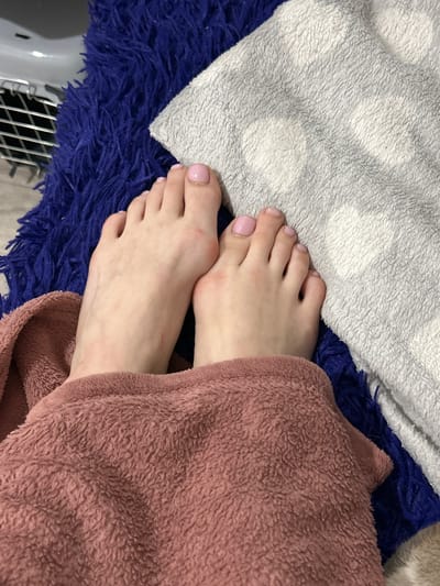 The image shows a close-up view of a person's feet, which are resting on a collection of blankets. One foot is more prominent, with toes painted in a light pink color. The feet are positioned on a deep blue, shaggy rug. Next to the rug, a light grey blanket with a subtle polka dot pattern is visible. Partially covering the feet and extending into the foreground is a soft, rose-colored blanket. The background elements suggest an indoor setting, possibly a home environment, with a metal grate and a glimpse of what might be a piece of furniture to the upper left. The scene is focused on the feet and the textures of the soft furnishings. There is no specific indication of the city of Melitopol, Ukraine, within the image itself.