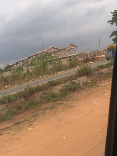 A low-angle shot captures a roadside scene with partially constructed buildings. The structures exhibit exposed frameworks, suggesting ongoing construction. Vegetation lines the road, separating it from a dirt area on the right. A blue truck is partially visible in the distance, driving down the road. The sky is overcast, with a dark cloud cover. The photograph appears to have been taken from a moving vehicle, as evidenced by the blurry foreground on the upper-right.