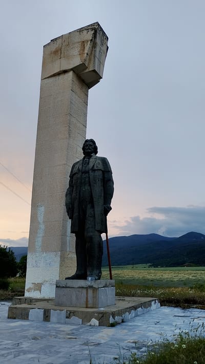 A weathered, dark bronze statue of a man in a suit and overcoat stands next to a tall, monolithic concrete monument in Kran, Bulgaria. The statue is positioned on a concrete pedestal, which is part of a larger, cracked marble-tiled plaza. The man in the statue holds a cane or staff. The monument is a light-colored, rough-hewn stone structure with geometric angles at its top, showing signs of decay and staining. The background features rolling hills and mountains under a twilight sky. A field with scattered wildflowers is visible between the monument and the hills. Power lines are visible in the distance to the left. The scene appears to be an outdoor memorial or monument site.