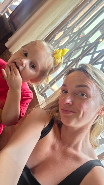 This media file is a selfie of a woman and a young child, likely mother and daughter, taken indoors. The woman, with blonde hair and fair skin, is in the foreground, looking towards the camera with a relaxed expression. She is wearing a black top. The child, also with blonde hair and fair skin, is behind and to the side of the woman, smiling and touching her mouth. The child is wearing a bright pink top and a yellow bow in her hair. The background shows a window with blinds or curtains and a metal grate structure, suggesting an interior space with natural light. The scene appears to be casual and intimate, capturing a moment between the two subjects. There is no visible text in the image. The lighting suggests it could be daytime. Given the provided location context of Guía de Isora, Spain, it's possible this photo was taken there.